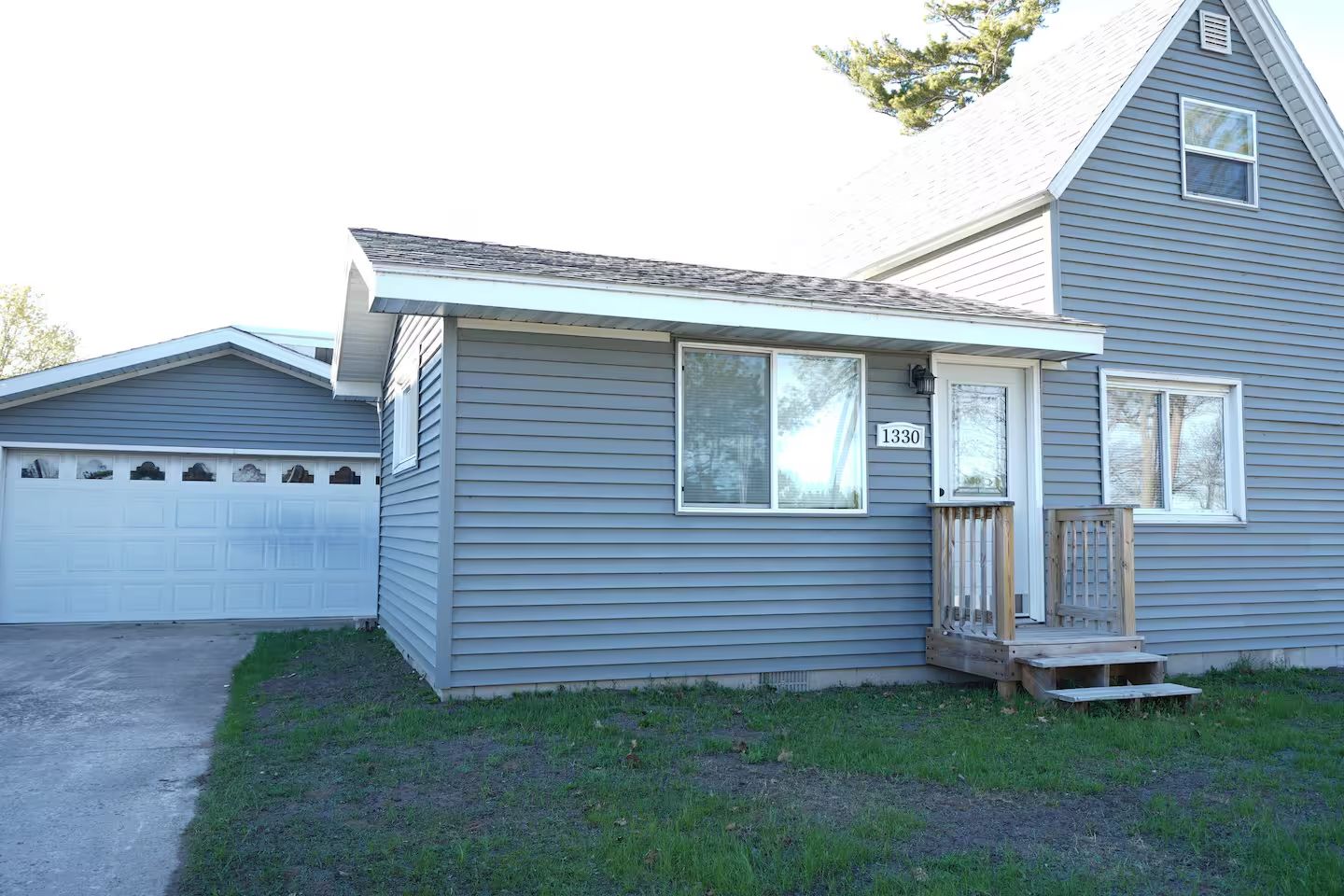 Gray house with white trim, small porch with steps, modern garage on the left, number 1330 on the wall. Surrounding lawn is partly sparse. Clear sky.