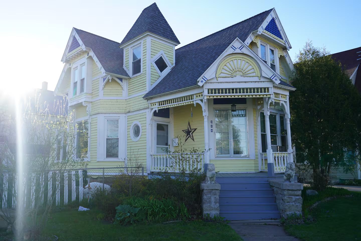 Victorian-style yellow house with ornate white trim and a steep roof. Sunlight softly highlights the garden and stone pathway, creating a calm ambiance.